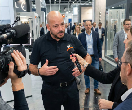 René Noël gibt ein Interview auf einer Industriemesse und spricht über Lockout/Tagout, Arbeitssicherheit und Safety-Kultur, während ein Kamerateam und Reporter ihn filmen.